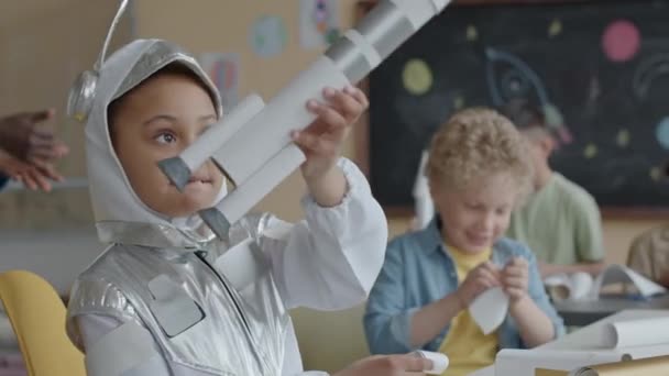 Little African American girl in astronaut costume playing with handmade paper rocket and then smiling on camera while sitting in classroom during lesson - Footage, Video