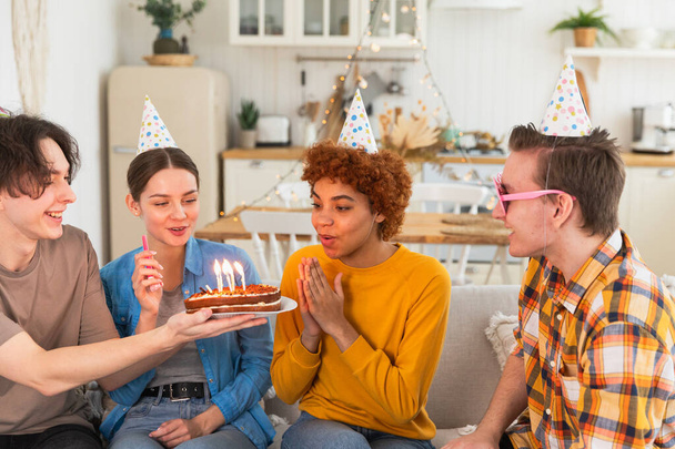 Doe een wens. Een vrouw met een pet die kaarsen uitblaast op verjaardagstaart. Gefeliciteerd met je verjaardag. Vriendengroep wenst meisje gefeliciteerd. Mensen die thuis hun verjaardag vieren met een feestje - Foto, afbeelding