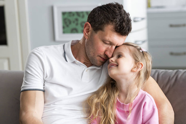 Eine kleine Tochter sitzt auf dem Sofa im Zimmer neben ihrem schlafenden Vater, der sie umarmt. Ein Mädchen schaut Papa an und lächelt süß. Das Konzept der glücklichen Familie, Kindheit und Vaterschaft. Nahaufnahme - Foto, Bild