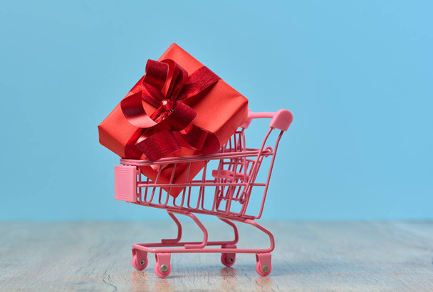 Ein in rotes Papier gewickeltes Geschenk in einem Miniatur-Einkaufswagen auf blauem Hintergrund - Foto, Bild