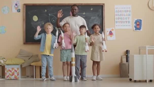 Group portrait of joyous African American teacher and little multi-ethnic kids holding paper rockets, smiling and waving on camera in school classroom - Footage, Video