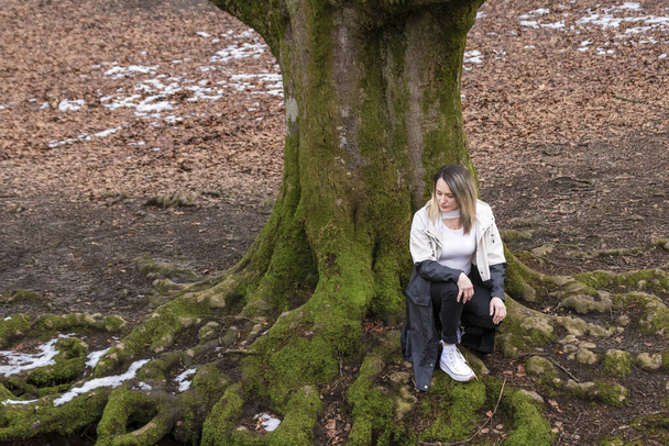 森の中の木の根の上に座っている女性,白いシャツ,黒いズボン,白い靴を着て - 写真・画像