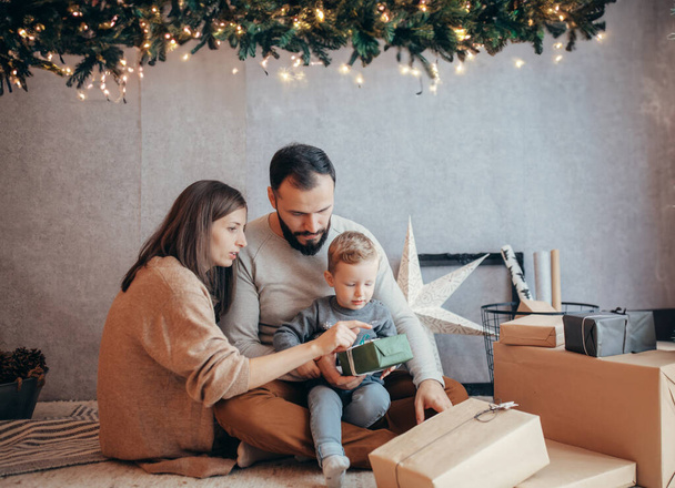 Περιτριγυρισμένο από μια μυριάδα από δώρα και λαμπερά φώτα σε ένα στούντιο στολισμένο με χριστουγεννιάτικα μοτίβα, μια νεαρή οικογένεια, το παιδί τους στο κέντρο, αποπνέει απόλαυση και γιορτινή διάθεση, αιχμαλωτίζοντας το πνεύμα της εποχής σε αυτό το ζωντανό σκηνικό - Φωτογραφία, εικόνα