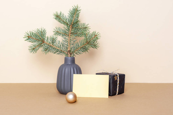 Seramik vazodaki ladin dalı, hediye kutusu ve bej masadaki boş kart. Asgari Noel konsepti. Ön görünüm, model. - Fotoğraf, Görsel