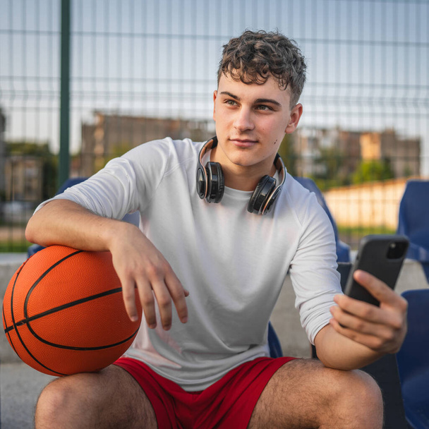 Кавказский подросток с помощью мобильного телефона смартфона на баскетбольной площадке - Фото, изображение