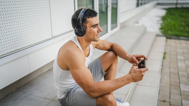 Dışarıdaki genç adamın portresi video oyunları oynamak için kulaklık ve akıllı telefon kullanıyor. - Fotoğraf, Görsel