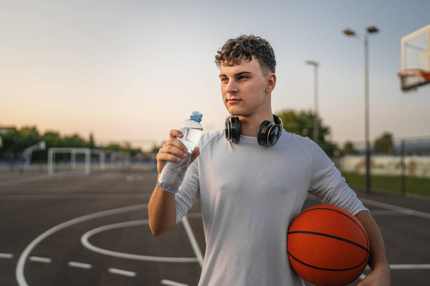 caucásico hombre adolescente abierto botella de plástico de agua al aire libre en día soleado beber mientras agujero baloncesto - Foto, Imagen