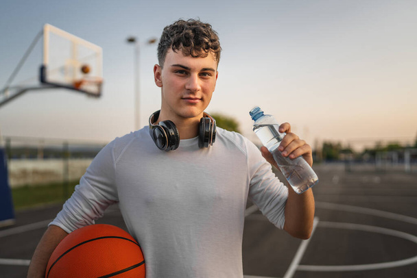 homme caucasien adolescent ouvert bouteille en plastique d'eau en plein air dans une boisson ensoleillée jour tandis que le basket-ball trou - Photo, image