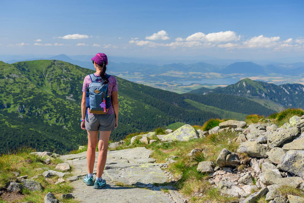 Polské Tatry, mladá žena v kraťasech a čepici s horským batohem stojí na horské stezce a dívá se na údolí s vrcholky hor za letního slunečného dne. - Fotografie, Obrázek