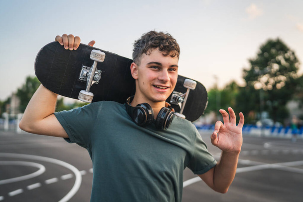 One man young adult caucasian teenager stand outdoor with skateboard on his shoulder and headphones posing at basketball court portrait happy confident wear shirt casual real person copy space - Photo, Image