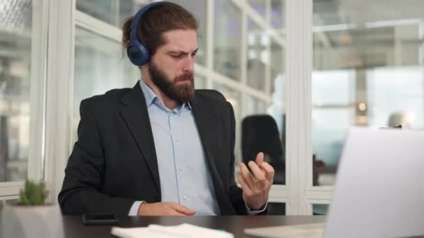 Vicces üzletember, aki vezeték nélküli kék fejhallgatót hord, gitározik, miközben laptoppal ül a modern irodában. Izgalmas férfi alkalmazott hallgat kedvenc zene és szórakozás beltérben. - Felvétel, videó