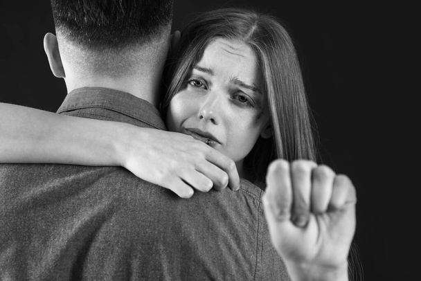 Bruised young woman with her husband showing fist on dark background. Domestic violence concept - Photo, Image