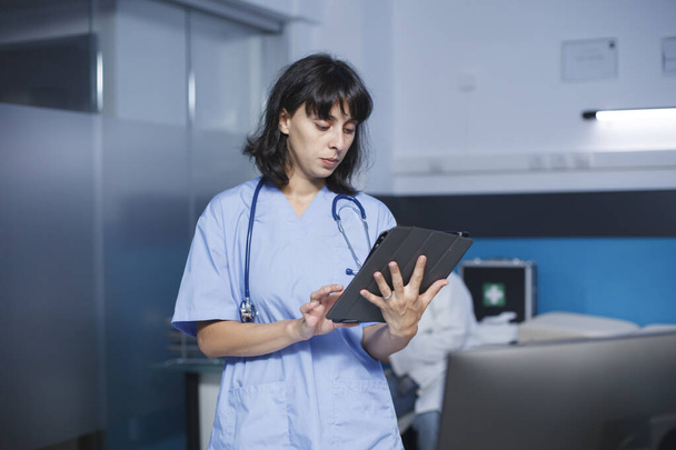 Szczegółowy obraz białej kobiety w niebieskim fartuchu stojącej w gabinecie kliniki, używającej tabletu do badań medycznych. Pielęgniarka wykorzystująca cyfrowe urządzenie do sprawdzania wizyt u pacjentów. - Zdjęcie, obraz