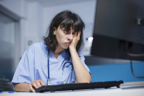 Close-up shot of a tired Caucasian woman with a stethoscope working on desktop PC in the clinic office. Selective focus of a worn out nurse in blue scrubs using a computer. Professional and detailed. - Photo, Image