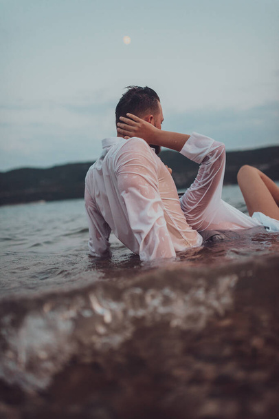 日没の静かな海の美しさの中で情熱的なキスを共有するロマンチックな若いカップル - 写真・画像