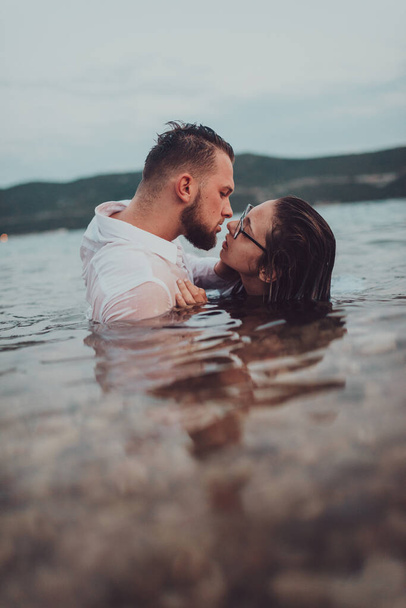 Una romántica pareja joven compartiendo un beso apasionado en medio de la serena belleza del océano al atardecer - Foto, imagen