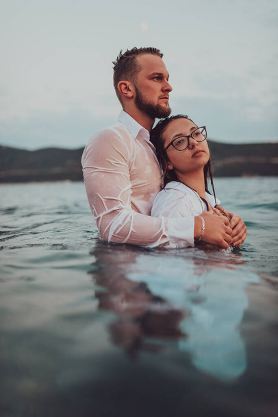 Una coppia simbolica ed emotivamente connessa si abbraccia teneramente sullo sfondo di un mare sereno, immerso nelle calde tonalità di un tramonto mozzafiato.  - Foto, immagini