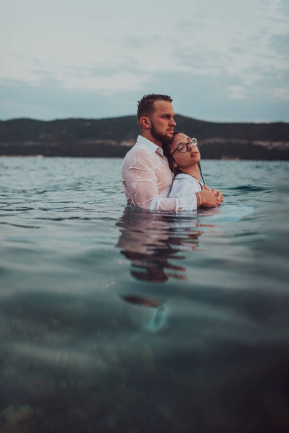 Um casal simbólico e emocionalmente ligado abraça ternamente o pano de fundo de um mar sereno, banhado nos tons quentes de um pôr-do-sol deslumbrante..  - Foto, Imagem
