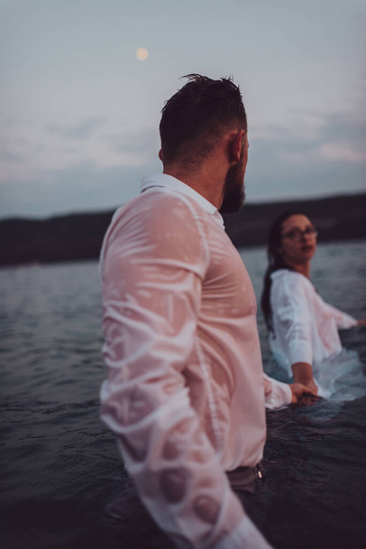 Un jeune couple, vêtu de vêtements, tient symboliquement la main tout en se tenant dans la mer pendant le coucher de soleil envoûtant, créant une scène pittoresque d'amour et d'unité dans le contexte de l'océan - Photo, image