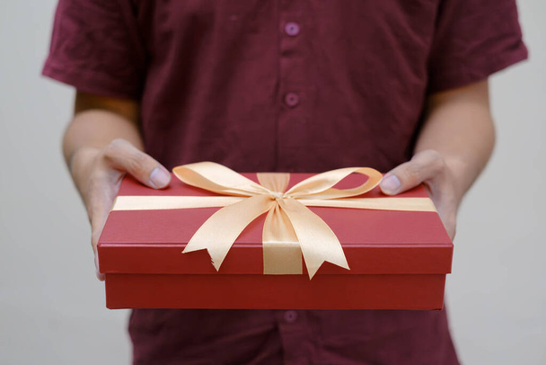 Αρσενικά χέρια κρατώντας ένα μικρό κόκκινο κουτί δώρου τυλιγμένο με χρυσή κορδέλα. Κοντινό πλάνο και εσωτερική λήψη. Επιλεκτική εστίαση. Χριστουγεννιάτικη ιδέα. - Φωτογραφία, εικόνα
