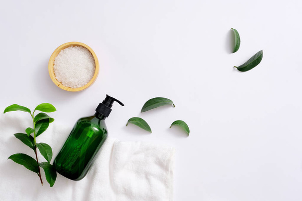 Salt is stored in a small wooden plate. The unlabeled shower gel bottle is placed on the towel along with fresh green tea leaves. Blank label for branding and advertising. - Photo, Image
