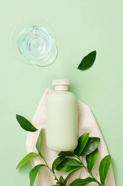 El té verde es un ingrediente popular en cosméticos. Una botella de gel de ducha sin etiqueta se coloca junto a una toalla de tela y hojas de té verde fresco, un vaso de agua sobre un fondo minimalista. - Foto, imagen