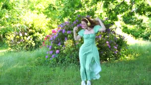 Retrato al aire libre de mujer pelirroja feliz en vestido verde y auriculares bailando en el parque público. Escuchar música en línea en cualquier lugar y ser tú mismo concepto - Imágenes, Vídeo