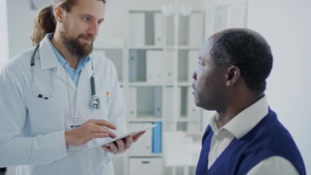 Inyección lateral media del practicante caucásico con tableta preguntando al paciente varón negro sobre las quejas durante la consulta de chequeo en el hospital - Metraje, vídeo