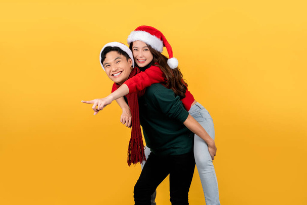 Sorridente coppia asiatica indossa maglioni e cappelli di Babbo Natale giocosamente posa a cavalluccio su uno sfondo giallo. Felice anno nuovo o concetto di vacanza di Natale. - Foto, immagini