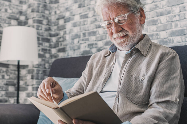 Headshot portrait close up of old happy and relaxed man sitting reading a book at home. Mature male person enjoying free time having fun indoor. - Foto, imagen
