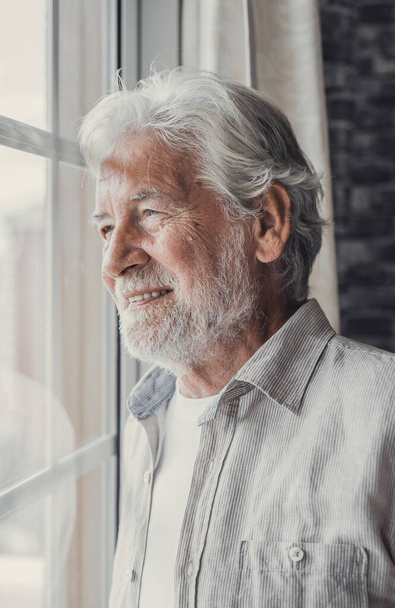Ευτυχισμένος στοχαστικός ηλικιωμένος άνδρας της δεκαετίας του '70 που κοιτάζει από το παράθυρο μακριά με ελπίδα, σκέφτεται την καλή υγεία, συνταξιοδότηση, ασφαλιστικές παροχές, ονειρεύεται το μέλλον. Ηλικιωμένοι συνταξιούχοι αναμένουν συνάντηση με την οικογένεια - Φωτογραφία, εικόνα