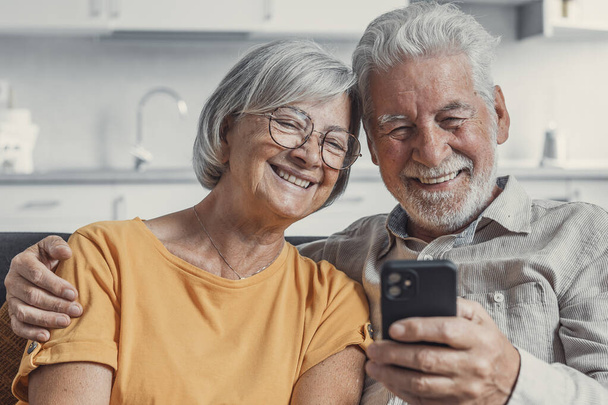 Szczęśliwa dojrzała Rodzina, żona i mąż za pomocą telefonu razem w domu, uśmiechając się w średnim wieku mężczyzna i kobieta siedzi na kanapie, za pomocą aplikacji urządzenia mobilnego, oglądanie wideo w sieci społecznej, surfowanie po Internecie - Zdjęcie, obraz