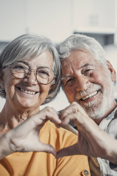 Közelkép portré boldog őszinte középkorú idős nyugdíjas család pár, hogy a szív gesztus ujjaival, kimutatva a szeretet, vagy őszinte érzelmek együtt zárt térben, nézi a kamera. - Fotó, kép