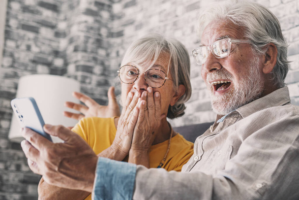 Happy older couple sit on couch staring at cellphone screen gesturing looking overjoyed, scream with joy, read fantastic news, get great commercial offer, pension raise. Success, achievement concept - Photo, image