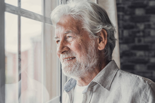 Onnellinen harkittuja vanhempi 70s mies katselee ulos ikkunasta pois toivoa, ajatellut hyvää terveyttä, eläkkeelle, vakuutus etuja, haaveilee tulevaisuudesta. Tapaamista perheen kanssa odottava iäkäs eläkeläinen - Valokuva, kuva