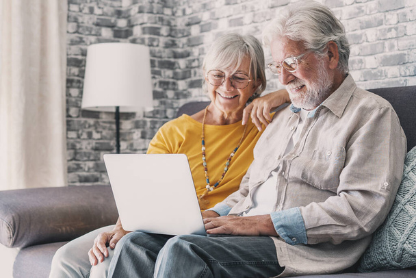 かなり高齢者70代の白髪のカップルは、リビングルームでソファの上に休んでラップノートパソコンの映画を見て笑顔で自由時間をお楽しみください,高齢者世代と現代の無線技術高度なユーザーコンセプト - 写真・画像