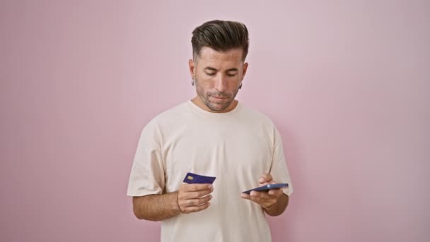 Huolestunut nuori latino mies seisoo vaaleanpunainen tausta, kamppailee online-maksu älypuhelimella. vakava kaveri järkyttynyt kirjoittaessaan luottokortin tiedot puhelimeen, eristetty hänen ongelmansa. - Materiaali, video
