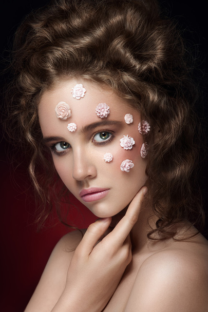 Romantic nude young beautiful girl with white flowers on her face and soft curls on dark background. Touching her face - Zdjęcie, obraz