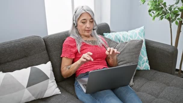 Schöne reife grauhaarige Frau zu Hause, die sich müde auf den Rücken streckt und nach der Arbeit am Online-Laptop auf dem gemütlichen Wohnzimmersofa die Kopfhörer abnimmt. - Filmmaterial, Video