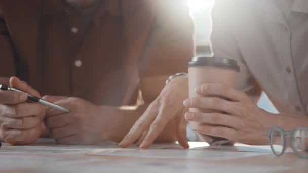 Midsection slowmo dwóch nierozpoznawalnych kolegów w luźnym ubraniu opierających się na stole i planujących projekt podczas przerwy kawowej w biurze w słoneczny dzień - Materiał filmowy, wideo