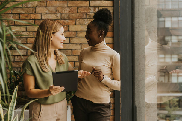 産業スタイルのオフィスの煉瓦の壁によって立っているデジタル タブレットが付いている2人のかなり若いビジネス 女性 - 写真・画像
