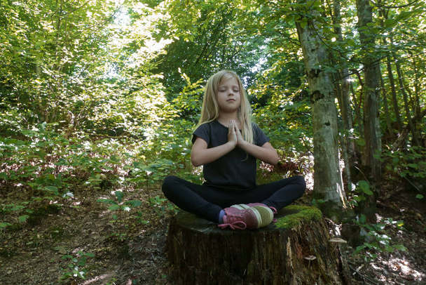 ツリー・スタンプの上に座っている 蓮の位置にいる小さな女の子。 森林瞑想のコンセプト。 平和,マインドフルネス,リラクゼーション. - 写真・画像