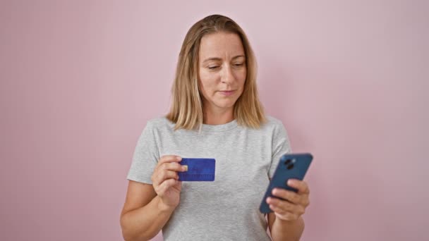 Boldog, fiatal szőke nő magabiztosan gépel fizetési üzenetet okostelefonon, tartja a hitelkártyáját és véletlenül áll. gyönyörű kifejezés rózsaszín elszigetelt háttér fal - Felvétel, videó