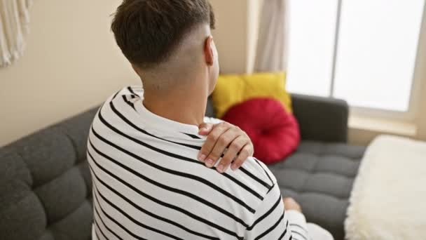 Hombre hispano joven preocupado sentado en el sofá en casa, sufriendo dolor de espalda grave, una realidad dolorosa en el interior. - Metraje, vídeo