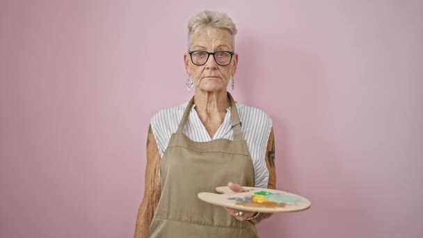 Femme artiste mature aux cheveux gris, avec pinceau et palette, peignant avec une grande précision sur un chevalet portable dans une classe d'art isolée et détendue au mur rose. - Séquence, vidéo