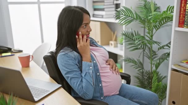 Successful young hispanic pregnant business woman, smiling while talking on her smartphone, subtly touching her belly, as she relaxes in her office room - Footage, Video