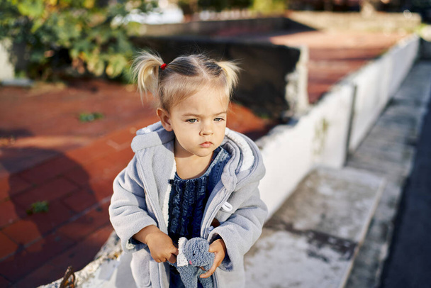 Mała dziewczynka z miękką zabawką w rękach siedzi na krawężniku w parku. Wysokiej jakości zdjęcie - Zdjęcie, obraz