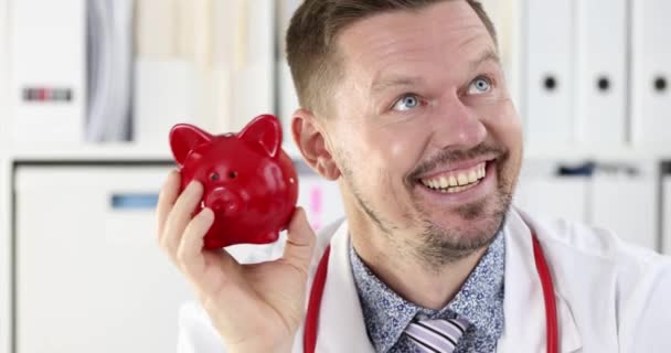Médico masculino bem sucedido sacudindo banco piggy vermelho perto de sua orelha filme 4k câmera lenta. Investimento no conceito de educação médica - Filmagem, Vídeo