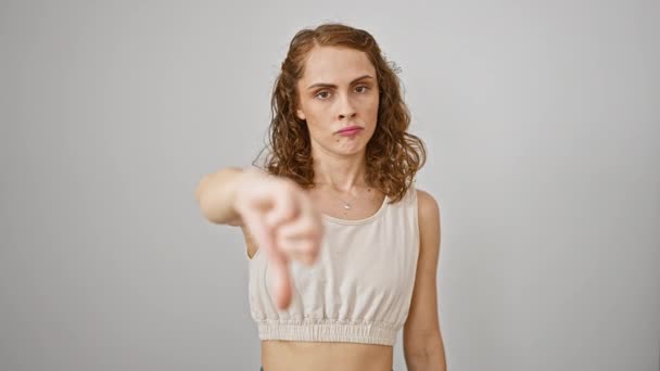 Приголомшлива молода жінка, стоячи ізольовано на білому тлі, зображує негативні великі пальці вниз жестом. її злий вираз обличчя і нещасний вираз демонструють схвалення - Кадри, відео