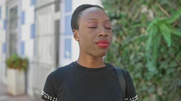 Cool, aantrekkelijk Afrikaans Amerikaanse vrouw staat ontspannen op een zonnige, stedelijke straat, belichamen levensstijl, met een ernstige uitdrukking en armen gekruist gebaar, tegen een casual achtergrond. - Video